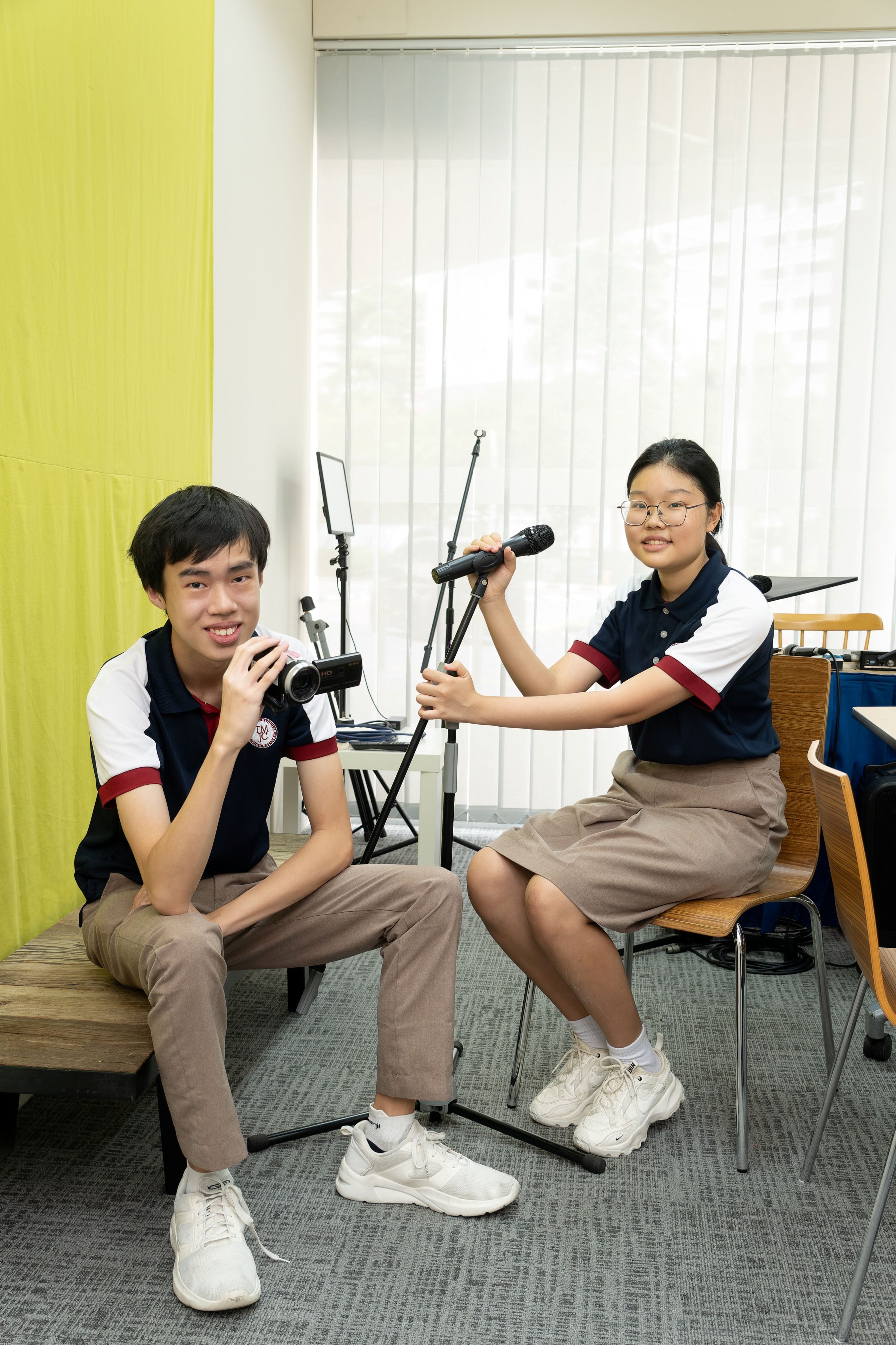 Two students in school uniforms sit in a media room. One holds a video camera, the other holds a microphone on a stand. Recording equipment is visible in the background.